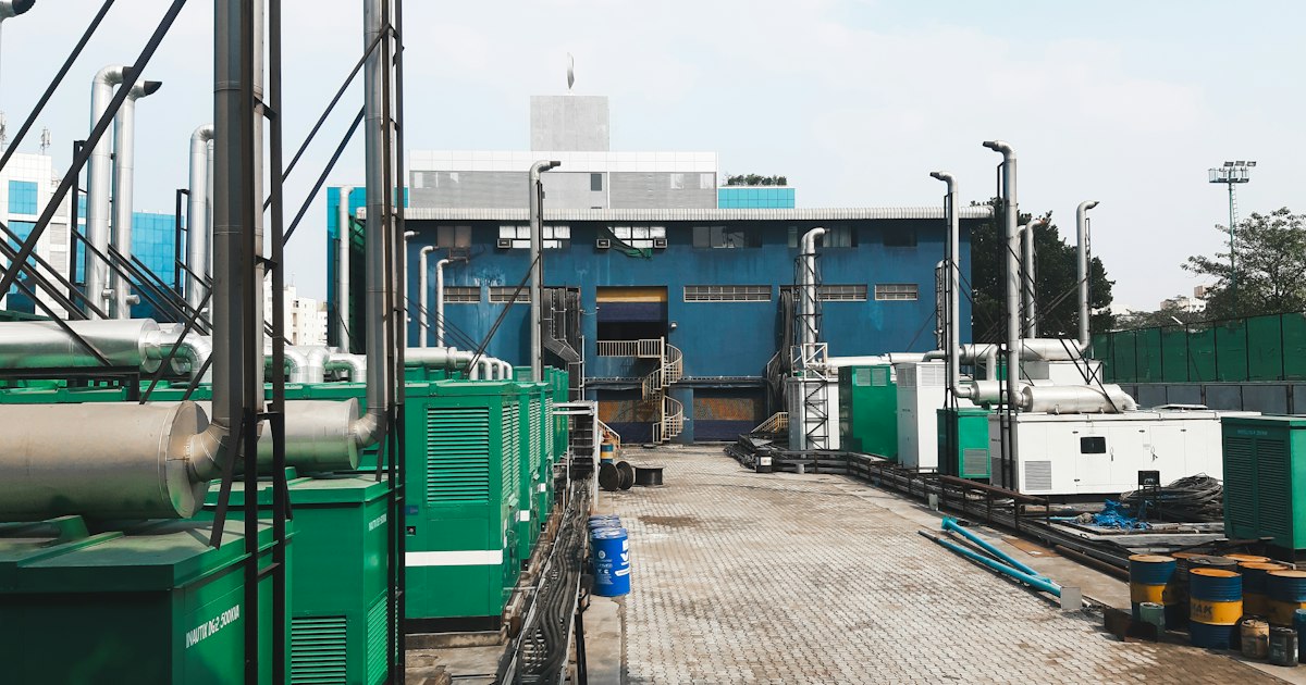 Row of green gas generator sets with exhaust stacks on a paved industrial pad beside oil drums