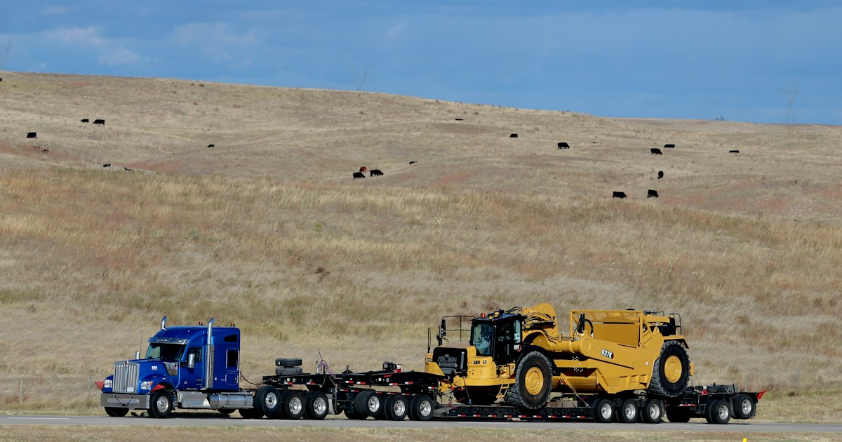Multi-axle lowboy trailer hauling heavy yellow earthmoving equipment along a rural highway